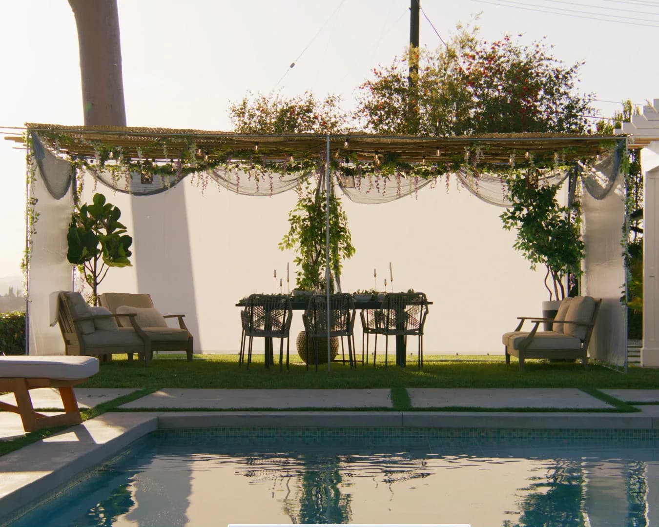 A sukkah in a sunlit backyard, its bamboo roof and white walls draped with greenery, the table set for a meal beside a pool.